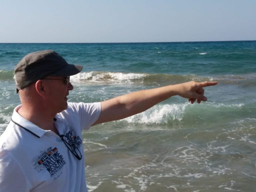 Mann zeigt auf das Meer, Wellen brechen am Strand im Hintergrund.
