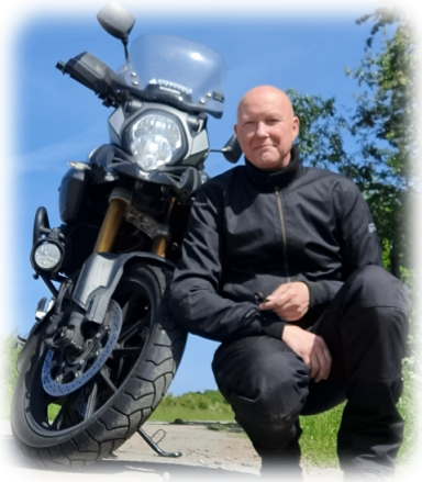 Mann in schwarzer Motorradbekleidung neben einem Motorrad auf einer Straße.